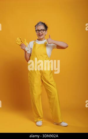 Die fröhliche ältere Frau, die einen gelben Overall trägt, lächelt und zeigt die Daumen hoch, während sie Gummihandschuhe hält, vor einem hellgelben Hintergrund. Stockfoto