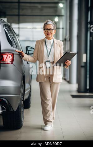 Eine selbstbewusste Frau in beigefarbenem Anzug steht neben einem eleganten SUV, hält ein Klemmbrett und einen Stift und demonstriert ihre Automobilkompetenz in einem modernen Händler Stockfoto