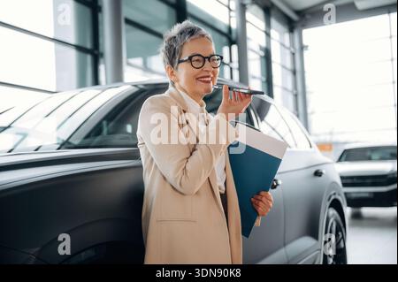 Professionelle Frau mit kurzen Haaren führt ein Telefongespräch, während sie eine blaue Mappe hält und in einem modernen Stil neben einem stilvollen Auto steht Stockfoto