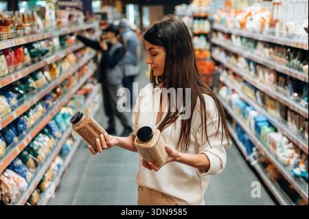 Eine junge Frau mit langen dunklen Haaren vergleicht nachdenklich zwei Gläser mit Gewürzen in einem Gang eines Lebensmittelgeschäfts, umgeben von Regalen mit verschiedensten Lebensmitteln Stockfoto