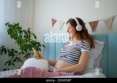 Schwangere Frau, die Musik oder Meditation über Kopfhörer genießt, während sie sich im Bett entspannt, das mütterliche Wohlbefinden und achtsames Zuhören für pränatale Entwicklung und Wohlbefinden annimmt Stockfoto