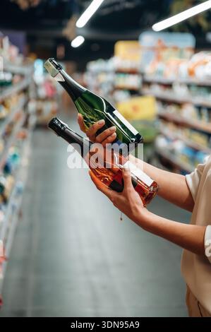 Die Hände halten zwei Flaschen Sekt, eine grüne und eine Bernsteinflasche, in einem Gang mit verschiedenen Produkten und Regalen, Highlight Stockfoto