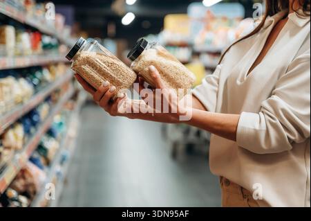 Weibliche Käuferin hält zwei Gläser Getreide in einem Lebensmittelgeschäft Gang und vergleicht Optionen, während sie von verschiedenen Lebensmitteln umgeben ist, um die Gesundheit zu betonen Stockfoto