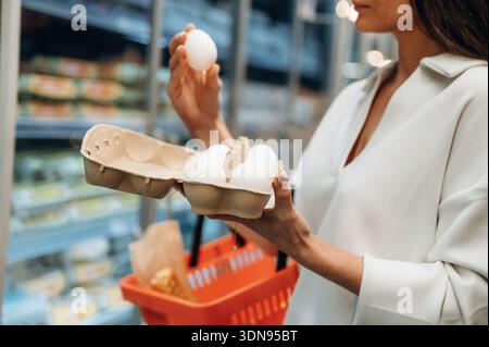 Die weibliche Käuferin hält einen Karton Eier in der einen Hand und ein Ei in der anderen Hand und prüft sorgfältig die Qualität, während sie von einer lebhaften Lebensmittelstraße umgeben ist Stockfoto