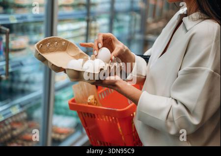 Weibliche Käuferinnen wählen sorgfältig frische Eier aus einem Karton aus, während sie einen roten Korb in einem Supermarkt halten, umgeben von bunten Produkten und gepflegt Stockfoto