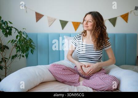 Schwangere Frau, die zu Hause Liebe und Pflege für ihr Baby zeigt, bequem gekleidet, während sie auf einem Bett sitzt, das mit einem Schwangerschaftskissen und Babyfellerei dekoriert ist, und das neue Leben vorwegnimmt Stockfoto