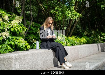 Eine entspannte Geschäftsfrau sitzt im Park und schreibt online per Smartphone SMS, während sie die Zeit im Freien genießt. Stockfoto
