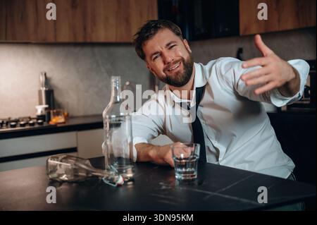 Kaukasier in weißem Hemd und Krawatte sitzt an einem Küchentisch, während er ein Glas Whiskey hält, mit einer leeren Flasche in der Nähe, Creatin Stockfoto