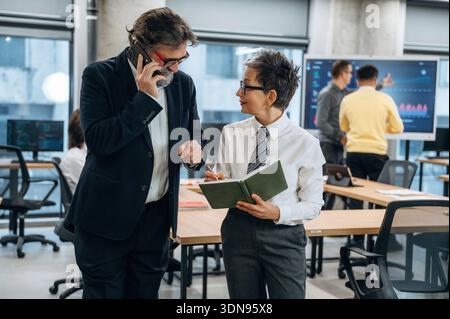 Zwei Geschäftsleute, einer auf einem Smartphone und der andere auf einem Notebook, arbeiten in einem modernen Bürobereich zusammen und heben den Tee hervor Stockfoto