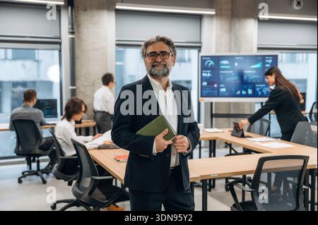 Ein selbstbewusster Geschäftsmann mit Brille und Bart steht in einem modernen Büro, umgeben von Kollegen, die an Computern arbeiten und Projekte diskutieren Stockfoto
