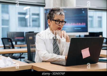 Eine reife Frau in formeller Kleidung arbeitet intensiv an einem Laptop in einem modernen Büro, mit digitalen Grafiken und Diagrammen auf Bildschirmen, Showcasin Stockfoto