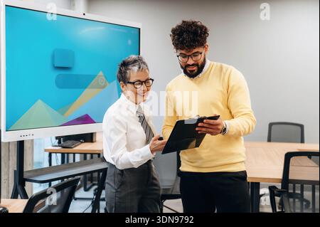 Zwei Geschäftsleute führen in einem modernen Büro eine gemeinsame Diskussion, prüfen Dokumente und tauschen Erkenntnisse aus, wobei Teammitglieder hervorgehoben werden Stockfoto