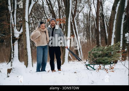 Eine Frau und ein Mann ziehen einen Weihnachtsbaum auf einem Schlitten durch einen verschneiten Wald, umgeben von hohen Bäumen und einer ruhigen Winterlandschaft Stockfoto