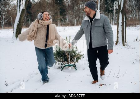 Glückliche Frau und Mann ziehen einen Weihnachtsbaum auf einem Schlitten durch eine verschneite Landschaft, umgeben von Bäumen, und zeigen einen fröhlichen Wintertag voller Freude Stockfoto