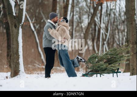 Afroamerikanische Männer und Frauen umarmen sich freudig in einem verschneiten Wald, gekleidet in gemütliche Winterkleidung, umgeben von Bäumen und einem Weihnachtsbaum, capt Stockfoto