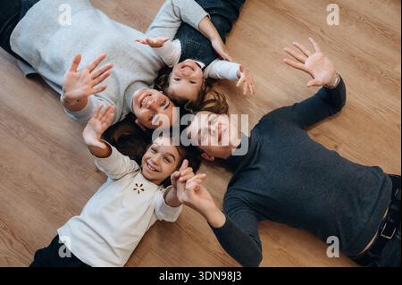 Vierköpfige Familie, einschließlich zwei Kindern, liegend auf einem Holzboden, lächelnd und mit den Händen winkend, schafft eine warme Atmosphäre der Freude und Verbundenheit, Showcasin Stockfoto