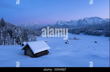 Schneebedeckte Holzhütte in einer gefrorenen alpinen Landschaft mit Bergen im Hintergrund während der blauen Stunde. Friedliche Winterlandschaft mit Neuschnee und Stockfoto