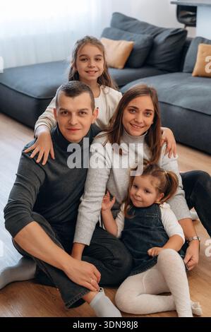 Vierköpfige Familie, einschließlich Eltern und zwei Töchtern, sitzt zusammen auf einem gemütlichen Wohnzimmerboden, umgeben von weichen Möbeln und natürlichem Licht Stockfoto