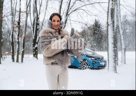 Stilvolle weibliche Figur in kuscheliger Winterkleidung, die in einer ruhigen Winterszene mit Bäumen und einem blauen Auto nea fröhlich einen Schal inmitten von Schneeflocken hält Stockfoto