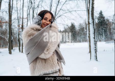 Frau mit dunklen Haaren, umhüllt in einen weichen Pelzmantel und Schal, lächelt in einem schneebedeckten Wald, umgeben von Bäumen, die Wärme und Glück im Winter verkörpern Stockfoto