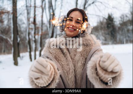 Glückliche Frau im warmen Wintermantel, die schneebedeckte Landschaft genießt, spielerisch mit funkelnden Lichtern interagiert und eine festliche und fröhliche Atmosphäre schafft Stockfoto