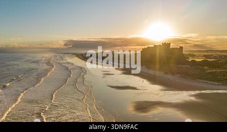 Das berühmte Bamburgh Castle an einem eiskalten Morgen bei Sonnenaufgang, neben dem schneebedeckten Strand in Northumberland, England, von Drohnen eingenommen. Stockfoto