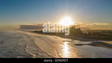 Das berühmte Bamburgh Castle an einem eiskalten Morgen bei Sonnenaufgang, neben dem schneebedeckten Strand in Northumberland, England, von Drohnen eingenommen. Stockfoto