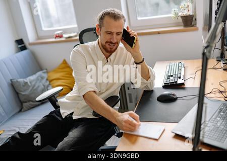 Ein Mann sitzt an einem Schreibtisch in einem gut beleuchteten Zimmer. Er hält ein Telefon am Ohr und macht Notizen auf einem Notizblock. Ein Computer und eine Tastatur befinden sich auf dem Schreibtisch. Stockfoto