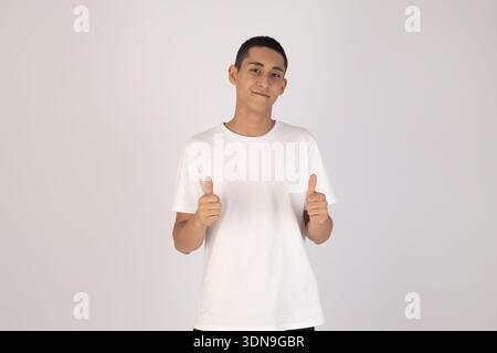 Glücklicher junger Mann, der den Daumen hoch auf weißem Hintergrund zeigt Stockfoto