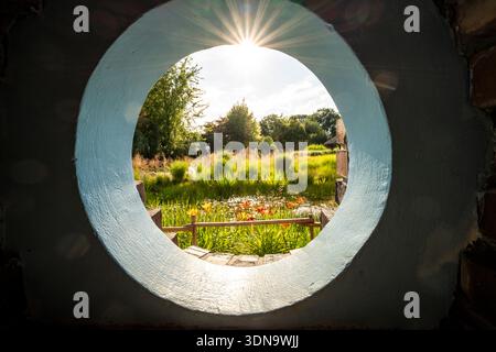 Blick durch ein rundes Fenster auf Ogrody Kapias in Polen, umgeben von bunten Blumen, üppigem Grün und ruhigen Gartenwegen in einer rustikalen, charmanten Umgebung Stockfoto