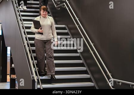 Weibliche Profis gehen eine stilvolle Treppe hinunter, halten einen Ordner, umgeben von moderner Architektur und einem eleganten Ambiente, das einen Dynami widerspiegelt Stockfoto