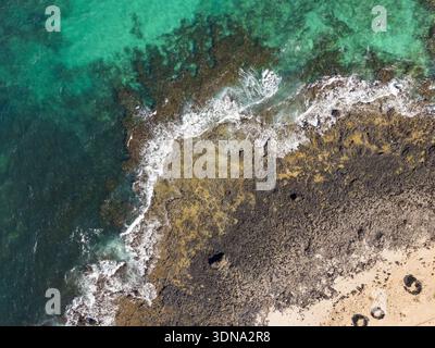 Aus der Vogelperspektive auf türkisfarbenes Wasser trifft auf die zerklüftete, dunkle vulkanische Küste, wo das Meer auf den Sandstrand trifft, Corralejo, Fuerteventura, Spanien. Stockfoto