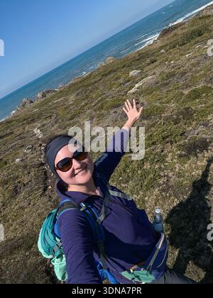 Lächelnder Alleinwanderer, der Selfie auf dem Küstenpfad macht, mit Meer und felsiger Küste im Hintergrund, Rucksack und Outdoor-Kleidung trägt. Reisen Stockfoto