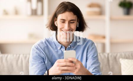 Junger Mann lächelt, während er tagsüber das Telefon zu Hause in einem hellen Wohnbereich benutzt Stockfoto