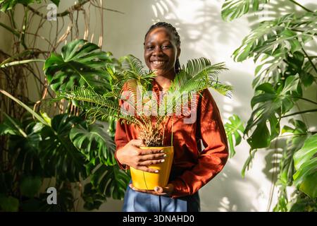 Die lächelnde afroamerikanische Frau hält Cycas im Topf, umgeben von üppig grünen tropischen Garten. Stockfoto