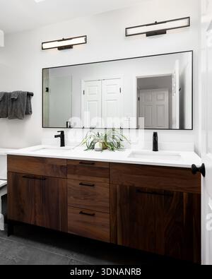 Ein moderner Kosmetikbereich im Bad verfügt über einen dunklen Holzschrank mit doppeltem Waschbecken mit weißen Arbeitsflächen und schwarzen Armaturen, beleuchtet durch zwei lineare Wandleuchten. Stockfoto