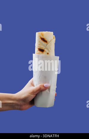 Hand hält eingewickeltes Shawarma in Papierhülle isoliert auf blauem Hintergrund Stockfoto