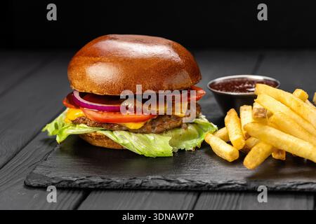 Rindfleisch-Cheeseburger mit Salat, Tomaten und Zwiebeln, serviert mit Pommes und Ketchup auf Schieferbrett Stockfoto