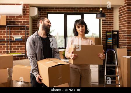 Glückliches junges Paar zieht in ein modernes Wohnhaus. Kaukasischer Mann und Frau stehen zusammen und halten Kisten mit vollgepackten Sachen, lächeln und nehmen neue Hausbesitzer auf. Stockfoto