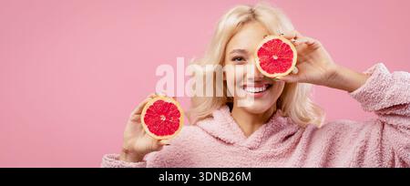 Die Frau hält Grapefruitscheiben nahe ihrem Gesicht und lächelt fröhlich vor rosa Hintergrund Stockfoto