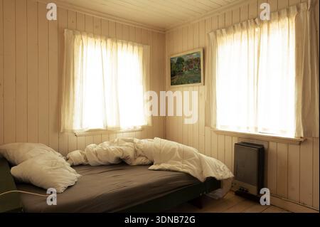 Ein unordentliches Bett mit weißen Bettdecken befindet sich in einem rustikalen, holzgetäfelten Schlafzimmer einer Ferienhütte, beleuchtet durch helles Morgenlicht von zwei Fenstern. Stockfoto