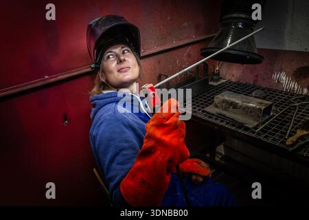 Schweißerin mit Schweißbrenner in einer industriellen Werkstatt, Schutzhandschuhe und Helm, Metallbearbeitungsumgebung. Stockfoto