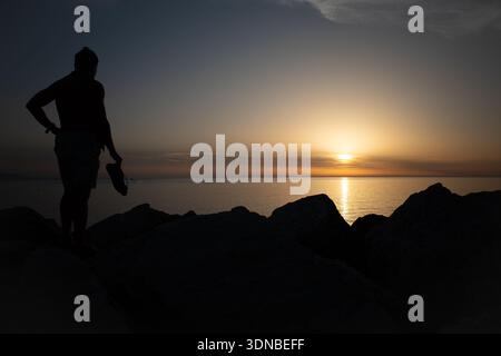 Silhouette eines Mannes, der bei Sonnenuntergang am felsigen Ufer steht, Schuhe hält und die goldene Sonne über dem ruhigen Horizont des Meeres reflektiert. Stockfoto