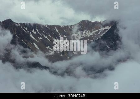 Schnee auf Berggipfeln ist sichtbar, während Wolken die Gipfel umgeben. Die Szene zeigt eine zerklüftete Landschaft mit felsigen Formationen und geringer Sicht durch Nebel. Stockfoto