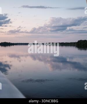 Sonnenuntergang über dem ruhigen Fluss mit pastellfarbenem Himmel. Ruhiger Fluss während der blauen Stunde: Der Himmel ist gefüllt mit weichen, strukturierten Wolken in Lavendel- und Blautönen. Stockfoto