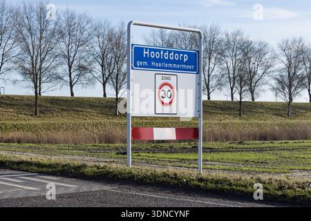 Blaues Straßenschild, das den Eingang zu Hoofddorp markiert, Gem. Haarlemmermeer, Niederlande, mit einem 30 km/h-Zonenschild an einem sonnigen Tag mit Bäumen dahinter. Stockfoto