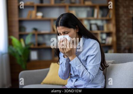 Junge Frau, die zu Hause auf dem Sofa sitzt und ihre Nase in ein Gewebe bläst, unwohl von saisonalen Allergien, Erkältung oder viralen Atemwegserkrankungen aussieht, sich müde und verstopft fühlt Stockfoto