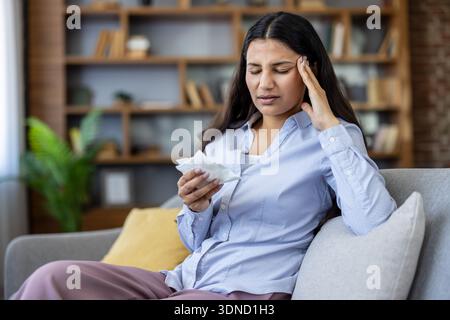 Junge Frau, die intensive Kopfschmerzen und Stress spürt, mit einer Hand ihren Kopf gewiegt und gleichzeitig ein Gewebe hält, zeigt Krankheit und Unwohlsein in einem häuslichen Umfeld Stockfoto