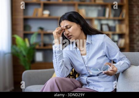 Frau, die schwere Migräne und Augenanstrengung erlitt, die Brille hielt und ihre Schläfen vor Schmerzen drückte, während sie auf einer Couch saß, erschöpft und gestresst von Überlastung zu Hause Stockfoto
