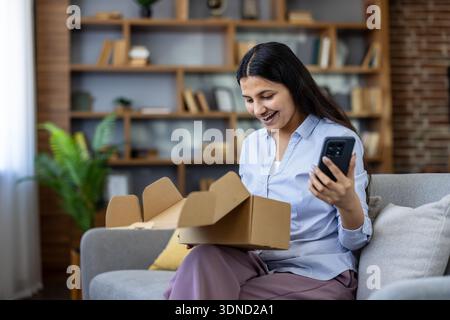 Glückliche Frau, die eine Online-Shopping-Lieferung auf dem Sofa auspackt, lächelt, während sie ihr Smartphone nach Bestelldetails sucht und in einer gemütlichen Wohnzimmerumgebung verfolgt Stockfoto